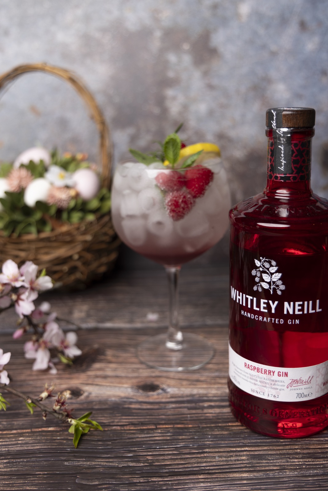 Eine Flasche des rot gefärbten Whitley Neill Raspberry Gin steht auf einer rustikalen Holzoberfläche neben einem großen Glas, das mit Eis und einer rosa Flüs…